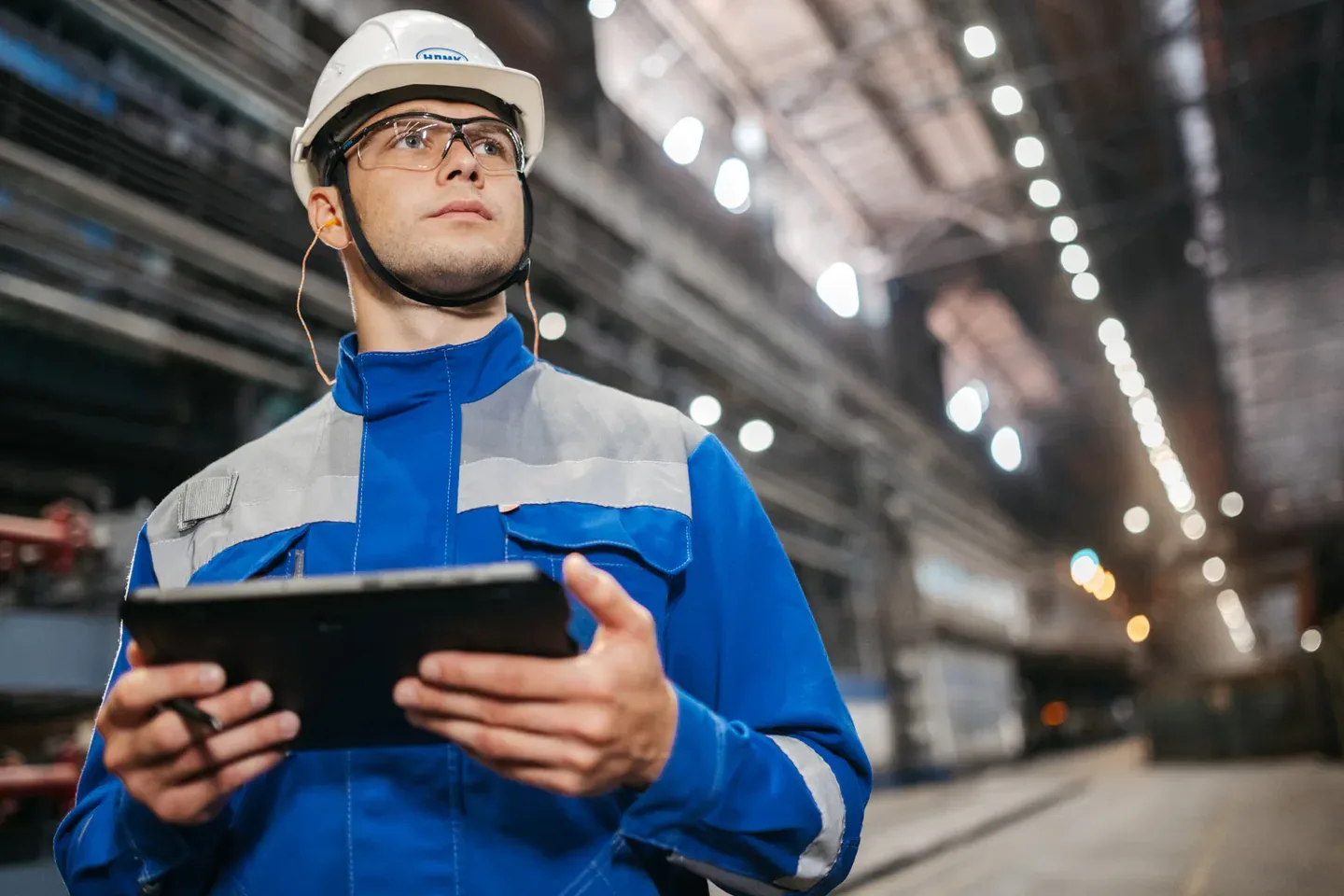 Production engineer in safety helmet reviewing order checklists on a tablet inside an industrial plant